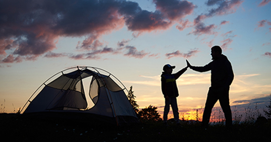 Silhouette of man and child hikers give each other a high five near the tent at dawn